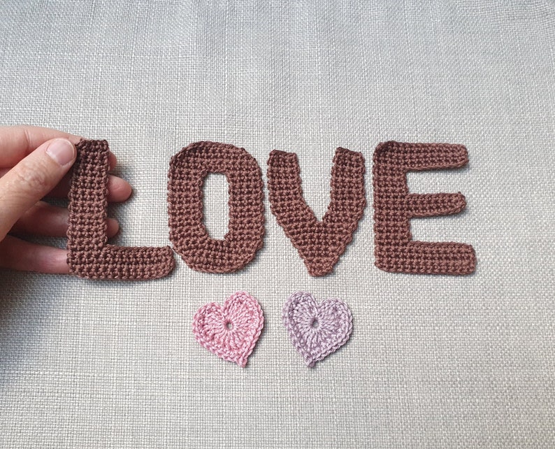 May include: Crocheted brown letters spelling "LOVE" with two small crocheted hearts, one pink and one lavender. The letters and hearts are handmade and placed on a neutral linen background. The letter "L" is being held by a hand.