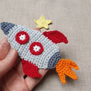 Könnte beinhalten: Gehäkelte Rakete in Grau, Rot und Orange mit blauer Spitze und roten Flügeln. Zwei rote und weiße Kreise sind zu sehen. Ein gelber Stern ist ebenfalls vorhanden.