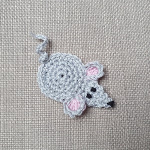 Op de afbeelding: Een gehaakte muisapplicatie in grijstinten, met roze binnenoren en zwarte ogen en neus. De muis heeft een gekrulde staart en is gemaakt van garen. De applicatie is waarschijnlijk handgemaakt en geschikt voor knutselwerk.