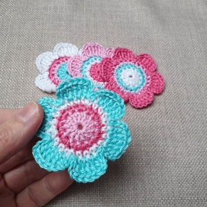 Könnte beinhalten: Fünf gehäkelte Blumen in den Farben Rosa, Weiß und Türkis. Die Blumen sind übereinander gelegt, wobei die türkisfarbene Blume im Vordergrund liegt.