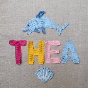 May include: Crocheted name "THEA" with a blue dolphin and a seashell. The letters are in pink, yellow, pink, and blue. The dolphin is light blue with darker blue accents. The seashell is light blue with white details.