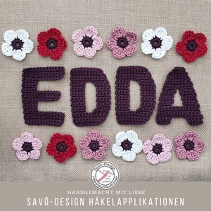 May include: A handmade crochet design featuring the name "EDDA" in dark purple yarn, surrounded by crochet flowers in white, pink, and red. The design includes the text "SAVO-Design H&auml;kelapplikationen" and "HANDGEMACHT MIT LIEBE".