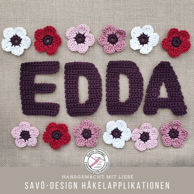 May include: A handmade crochet design featuring the name "EDDA" in dark purple yarn, surrounded by crochet flowers in white, pink, and red. The design includes the text "SAVO-Design H&auml;kelapplikationen" and "HANDGEMACHT MIT LIEBE".