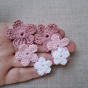 Könnte beinhalten: Eine Sammlung handgefertigter Häkelblumen in verschiedenen Rosa- und Weißtönen. Die Blumen haben ein fünfblättriges Design und sind in einer Gruppe angeordnet. Das Garn scheint weich und die Blumen sind klein.