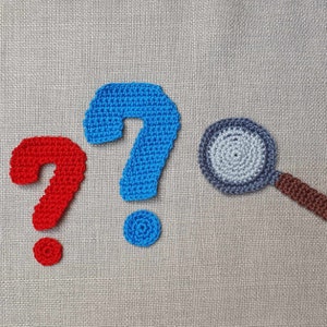 May include: Two blue crocheted question marks and one red crocheted question mark with a grey crocheted magnifying glass with a brown handle.