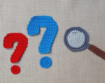 May include: A red crocheted question mark, a blue crocheted question mark, and a crocheted magnifying glass with a light grey lens and a brown handle. Each item is crafted with visible yarn stitches, creating a textured and dimensional appearance.