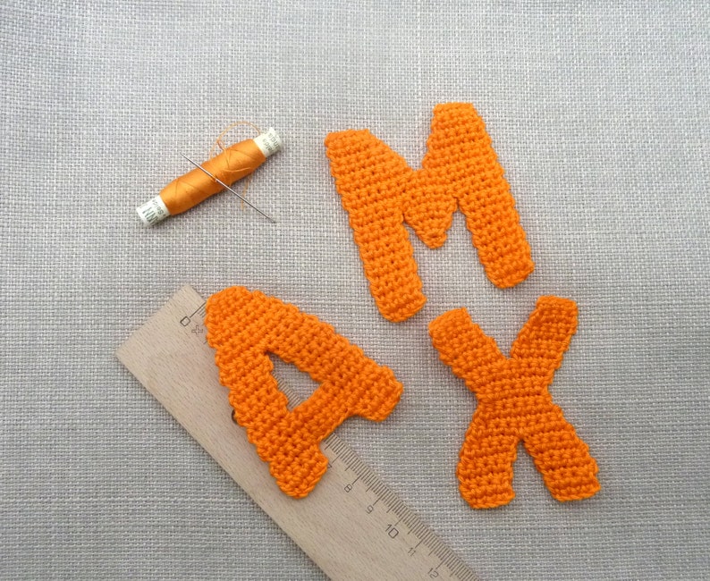 May include: Orange crocheted letters A, M, and X, along with a spool of orange thread and a needle. A wooden ruler is also present. The items are arranged on a gray textured surface.