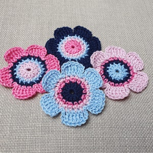 May include: Four crocheted flowers in pink, blue and navy blue. The flowers are arranged in a cluster on a beige fabric background.