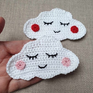 Könnte beinhalten: Zwei weiße gehäkelte Wolken mit lächelnden Gesichtern. Eine Wolke hat rote Wangen und die andere hat rosa Wangen.