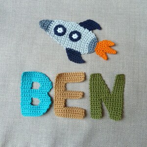May include: Crocheted rocket ship with blue, gray, and orange accents, and the name "BEN" spelled out in individual crocheted letters. The letters are in turquoise, tan, and green. The rocket has two white circles.
