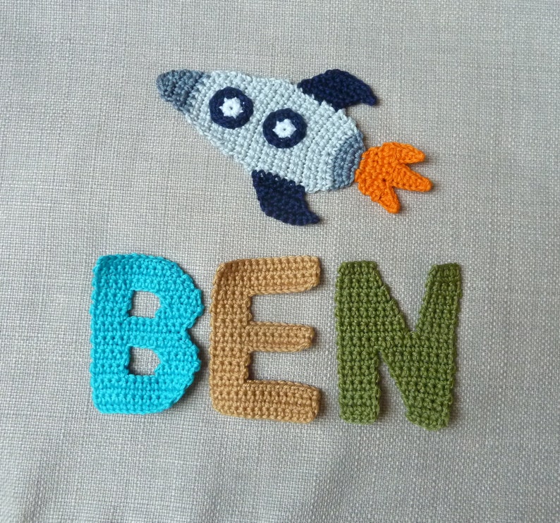 May include: Crocheted rocket ship with blue, gray, and orange accents, and the name "BEN" spelled out in individual crocheted letters. The letters are in turquoise, tan, and green. The rocket has two white circles.