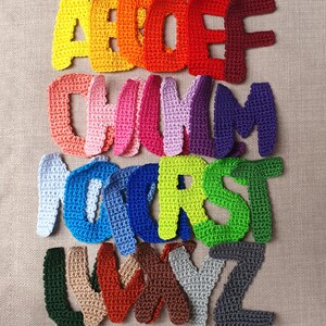 May include: A collection of colorful, crocheted letters of the alphabet. The letters are in various shades of yellow, orange, red, pink, purple, blue, green, brown, gray, and beige. The letters are arranged in rows on a neutral background.