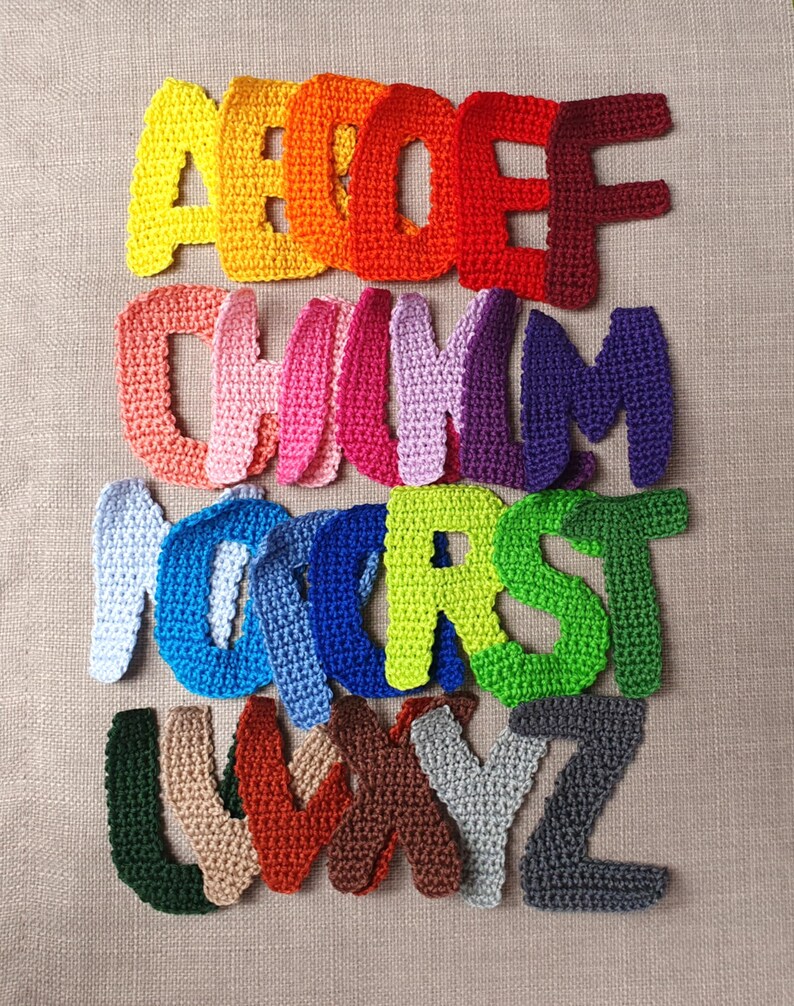 May include: A collection of colorful, crocheted letters of the alphabet. The letters are in various shades of yellow, orange, red, pink, purple, blue, green, brown, gray, and beige. The letters are arranged in rows on a neutral background.