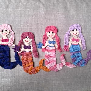 May include: Four crocheted mermaid ornaments with pink, purple, and orange tails. Each mermaid has a pink, purple, or white top and a smiling face.