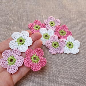 Könnte beinhalten: Eine Sammlung kleiner, fünfblättriger Häkelblumen in den Farben Weiß, Hellrosa und Dunkelrosa. Jede Blume hat eine leuchtend lindgrüne Mitte. Diese handgefertigten Blumenapplikationen sind mit einem einheitlichen kreisförmigen Muster gestaltet.