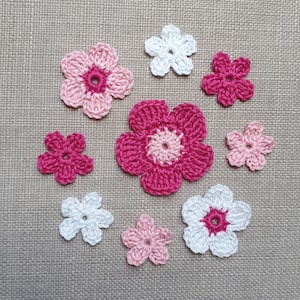 Può includere: Una collezione di nove fiori all'uncinetto rosa e bianchi. I fiori sono disposti in cerchio su uno sfondo di tessuto beige.