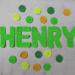 May include: A handmade crochet design spelling out the name "HENRY" in bright green yarn. The letters are surrounded by small, circular crochet embellishments in shades of green and gold, set against a gray fabric background.