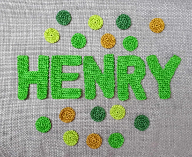 May include: A handmade crochet design spelling out the name "HENRY" in bright green yarn. The letters are surrounded by small, circular crochet embellishments in shades of green and gold, set against a gray fabric background.