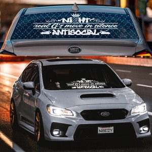 May include: A gray Subaru car with a white decal on the rear window that reads "NIGHT real G's move in silence ANTISOCIAL". The decal is surrounded by a black and white pattern. The car is parked on a street.