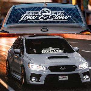May include: A white Subaru WRX with a black and white "Low & Slow" car window decal. The decal features a speedometer graphic and the text "Life is Better Low & Slow" in Japanese.
