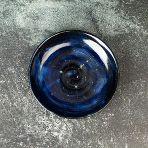 May include: A round, ceramic plate with a deep blue, swirling galaxy design. The plate features a constellation pattern with white and yellow star accents. The plate's rim is dark blue, and the center is a deep, dark blue.