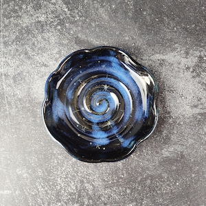 May include: A handmade ceramic dish with a wavy rim. The dish features a dark blue and black glaze with a spiral design in the center, and small white star-like accents. The dish is approximately 6 inches in diameter.