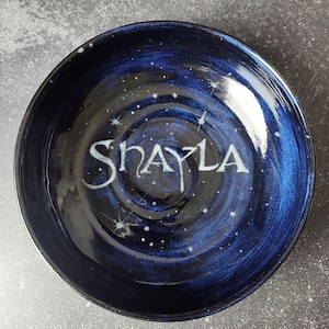 May include: A round ceramic bowl with a deep blue and black swirled glaze finish. The interior features a hand-painted white starfield pattern and the name "SHAYLA" written in a stylized, elegant white font across the center of the bowl.