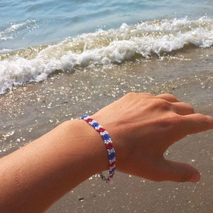 May include: A beaded bracelet with a pattern of red, white, and blue beads. The bracelet is on a person's wrist, with a beach and ocean in the background.