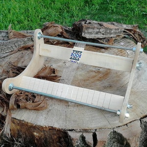 May include: A wooden loom with a beaded design in progress. The loom is made of natural wood and has a metal rod for weaving. The loom is sitting on a tree stump.