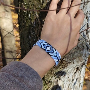 May include: A hand wearing a blue, white, and black beaded bracelet with a geometric pattern. The bracelet is on a person's wrist, and the hand is touching a tree trunk.