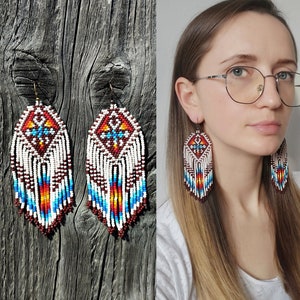 May include: A pair of long, beaded earrings with a white, red, blue and brown geometric pattern. The earrings are hanging from a wooden surface.