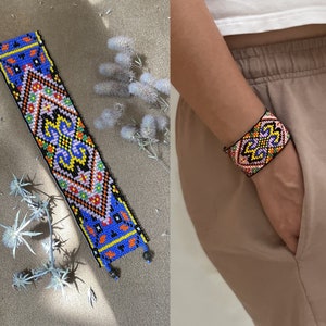 Könnte beinhalten: Ein farbenfrohes Armband aus Perlen mit einem geometrischen Muster in Blautönen, Rosa, Gelb und Schwarz. Das Armband besteht aus kleinen Perlen und hat ein gewebtes Design.