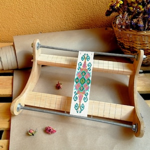 May include: A wooden bead loom with a colorful beaded design in progress. The loom has two levels and is made of light wood. The beaded design is pink, green, and blue.