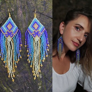 Könnte beinhalten: Ein Paar lange, geperlte Ohrringe mit einem geometrischen Muster in Blautönen, Lila und Gold. Die Ohrringe haben einen Fransen aus Perlen, der vom geometrischen Muster herabhängt.