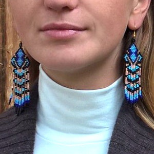 May include: A woman wearing a brown cardigan and a white turtleneck sweater with long blue and black beaded earrings.