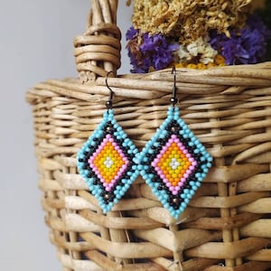 Könnte beinhalten: Ein Paar rautenförmige Ohrringe aus Perlen. Die Ohrringe haben ein zentrales Design aus gelben, rosa und schwarzen Perlen, umgeben von einem hellblauen Rand. Sie hängen an dunklen Metallhaken. Im Hintergrund befinden sich ein geflochtener Korb und Trockenblumen.
