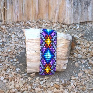 Puede incluir: Un brazalete de cuentas de colores con un patr&oacute;n geom&eacute;trico en tonos de morado, azul, amarillo y rojo. El brazalete est&aacute; sobre un trozo de madera.