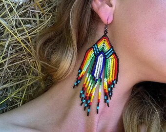 May include: A pair of long, colorful beaded earrings with a geometric design. The earrings are made of small, colorful beads in shades of red, orange, yellow, green, blue, and black. The earrings are hanging from a silver hook.