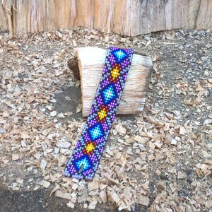 Puede incluir: Un brazalete de cuentas de colores con un patr&oacute;n geom&eacute;trico en tonos de morado, azul, amarillo y rojo. El brazalete est&aacute; sobre un trozo de madera.