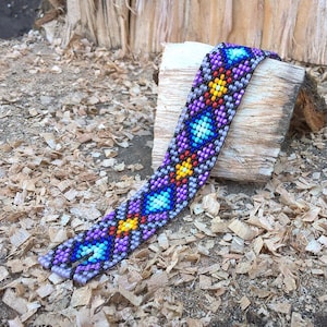 May include: A colourful beaded bracelet with a geometric pattern in shades of purple, blue, yellow, and red. The bracelet is made of small beads and is woven together in an intricate design.