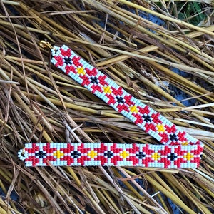 Könnte beinhalten: Ein rotes, weißes, schwarzes und gelbes Perlenarmband mit geometrischem Muster. Das Armband liegt auf einem Bett aus getrocknetem Stroh.