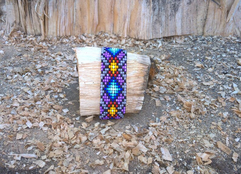 Puede incluir: Un brazalete de cuentas de colores con un patr&oacute;n geom&eacute;trico en tonos de morado, azul, amarillo y rojo. El brazalete est&aacute; sobre un trozo de madera.