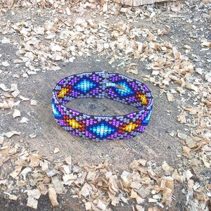Puede incluir: Un brazalete de cuentas coloridas con un patr&oacute;n geom&eacute;trico en tonos de morado, azul, amarillo y rojo. El brazalete est&aacute; hecho de peque&ntilde;as cuentas redondas y est&aacute; tejido en un patr&oacute;n apretado e intrincado.