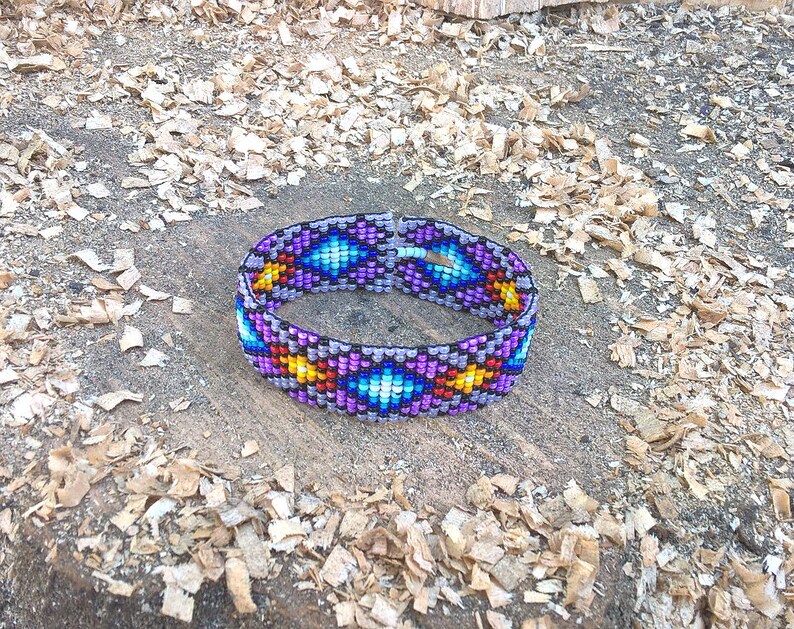 Puede incluir: Un brazalete de cuentas coloridas con un patr&oacute;n geom&eacute;trico en tonos de morado, azul, amarillo y rojo. El brazalete est&aacute; hecho de peque&ntilde;as cuentas redondas y est&aacute; tejido en un patr&oacute;n apretado e intrincado.