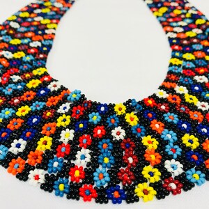 May include: A multi-colored beaded necklace with a floral pattern. The necklace is made of small, round beads in shades of red, orange, yellow, blue, green, and white. The beads are arranged in a repeating pattern of flowers.