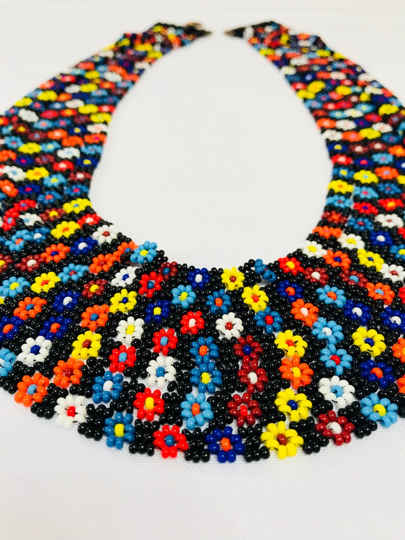 May include: A multi-colored beaded necklace with a floral pattern. The necklace is made of small, round beads in shades of red, orange, yellow, blue, green, and white. The beads are arranged in a repeating pattern of flowers.