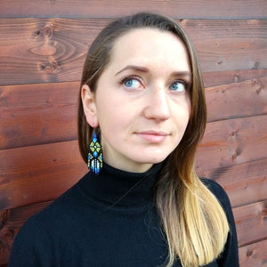 May include: A woman with long blonde hair wearing a black mock neck shirt and beaded earrings with blue, yellow, and white geometric patterns.