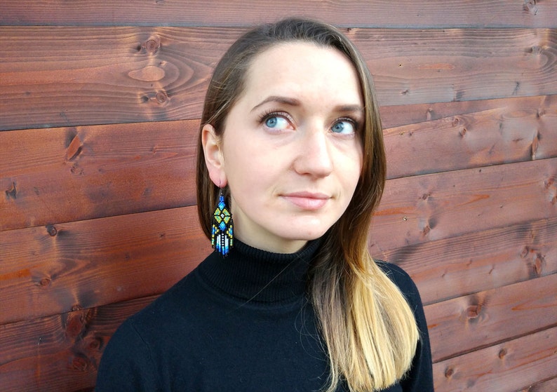 May include: A woman with long blonde hair wearing a black mock neck shirt and beaded earrings with blue, yellow, and white geometric patterns.