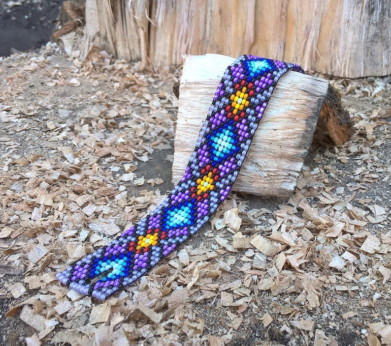 Puede incluir: Un brazalete de cuentas coloridas con un patr&oacute;n geom&eacute;trico en tonos de morado, azul, amarillo y rojo. El brazalete est&aacute; hecho de cuentas peque&ntilde;as y est&aacute; tejido en un dise&ntilde;o intrincado.