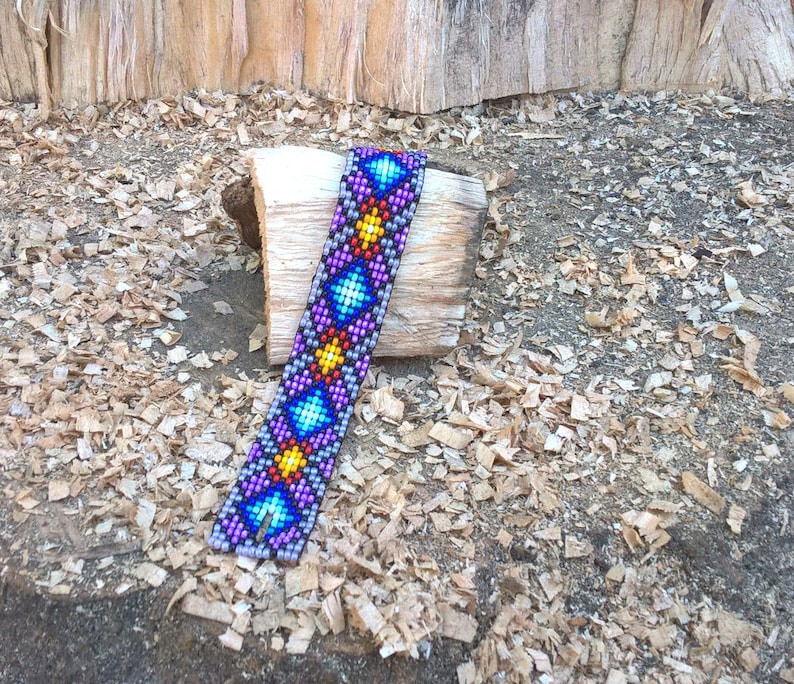 Puede incluir: Un brazalete de cuentas de colores con un patr&oacute;n geom&eacute;trico en tonos de morado, azul, amarillo y rojo. El brazalete est&aacute; sobre un trozo de madera.